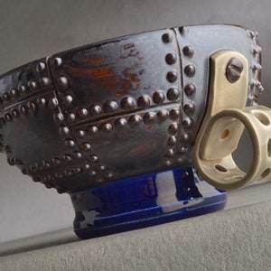 Può includere: Una ciotola in ceramica marrone con una base blu e un manico in metallo. La ciotola è decorata con rivetti e ha un'estetica steampunk.