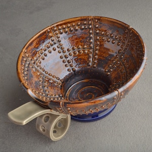 Puede incluir: Un cuenco de cerámica marrón con una superficie texturizada y una base azul. El cuenco está sobre un soporte con ruedas y un asa.