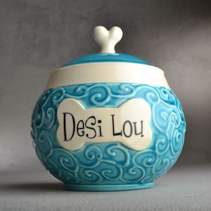 Pode incluir: Um pote de comida para animais de cerâmica azul e branco com uma tampa em forma de coração. O pote tem um design em forma de osso com o nome "Desi Lou" escrito nele.