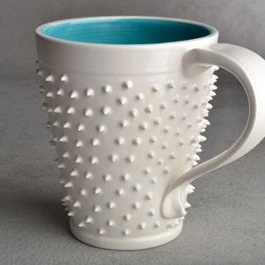 Könnte beinhalten: Weiße Keramiktasse mit türkisblauem Innenraum und einer strukturierten Außenseite mit kleinen, erhabenen Noppen. Die Tasse hat einen Henkel.
