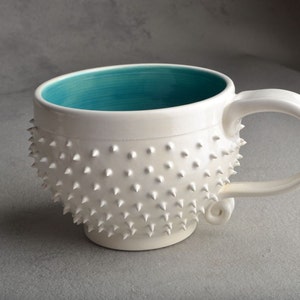 Peut inclure: Une tasse en céramique blanche avec un intérieur bleu turquoise et un extérieur texturé avec de petites pointes blanches. La tasse a une forme arrondie et une anse.