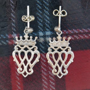 May include: A pair of silver-toned drop earrings. Each earring features a crown design above an openwork heart-shaped pendant. The earrings are displayed against a plaid background with red, black, and white colors.