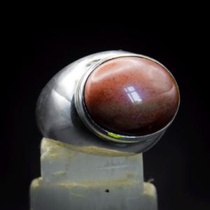 May include: A polished silver ring featuring a large, smooth, oval-shaped cabochon stone in shades of deep reddish-brown and muted pink. The metal band is thick and rounded, providing a solid frame for the prominent, glossy, multi-toned gemstone.