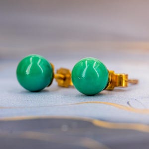 May include: A pair of stud earrings with bright, round, turquoise-coloured stones. The earrings have gold-coloured posts and backings. The background is a soft, blurred gradient of grey and gold.
