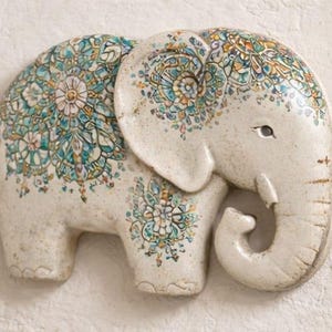 Op de afbeelding: Een crèmekleurig keramieken olifantenbeeldje met gedetailleerde turquoise, blauwe en gouden bloemmotieven. De olifant heeft een gekrulde slurf en grote oren.