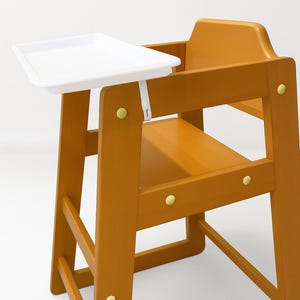 Puede incluir: Una trona de madera de color naranja cálido, con una bandeja blanca. La silla está fijada con fijaciones de color latón. La bandeja rectangular se asegura con una correa blanca. La silla tiene respaldo y reposapiés.