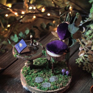 Miniature fairy garden set. Chair made with brown branches and green leaves and small purple velvet cushions. Decorated with green moss. Miniature glass table decorated with mushrooms and green moss with acorn teacup, candle and mini book.