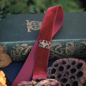 May include: A burgundy velvet ribbon bookmark with a silver-toned decorative clasp. The bookmark is draped over a vintage book with gold lettering and floral designs. A dried rose and seed pod are in the foreground.