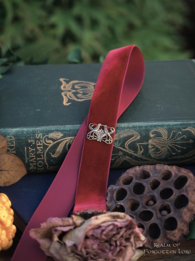 May include: A burgundy velvet ribbon bookmark with a silver-toned decorative clasp. The bookmark is draped over a vintage book with gold lettering and floral designs. A dried rose and seed pod are in the foreground.