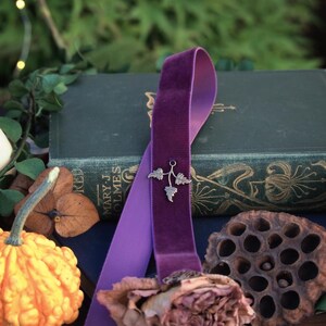 May include: A purple velvet ribbon bookmark with a silver leaf charm, draped over a vintage book. The bookmark rests near a dried rose, a decorative gourd, and a lotus pod. The background features greenery and the text "Realm of Forgotten Lore."