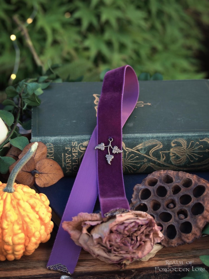 May include: A purple velvet ribbon bookmark with a silver leaf charm, draped over a vintage book. The bookmark rests near a dried rose, a decorative gourd, and a lotus pod. The background features greenery and the text "Realm of Forgotten Lore."