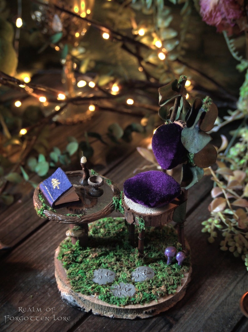 Miniature fairy garden set. Chair made with brown branches and green leaves and small purple velvet cushions. Decorated with green moss. Miniature glass table decorated with mushrooms and green moss with acorn teacup, candle and mini book.