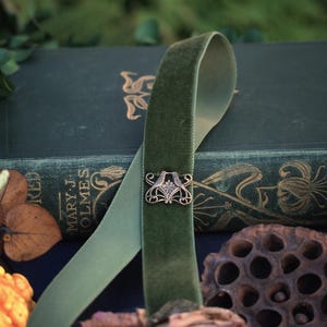 May include: A sage green velvet ribbon choker with an ornate silver-toned clasp. The ribbon is draped over a vintage book with gold lettering and floral designs. The image has a rustic, antique aesthetic.
