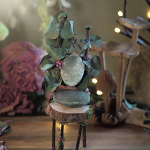 Miniature fairy chair made with brown branches and green leaves and small light green velvet cushions. Decorated with Dried pink flowers and green moss.