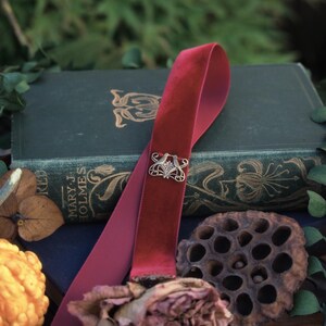 May include: A burgundy velvet bookmark with a silver filigree clasp rests on a vintage book. The bookmark is draped over the book, with a dried rose and other natural elements in the foreground. The text "Realm of Forgotten Lore" is visible.