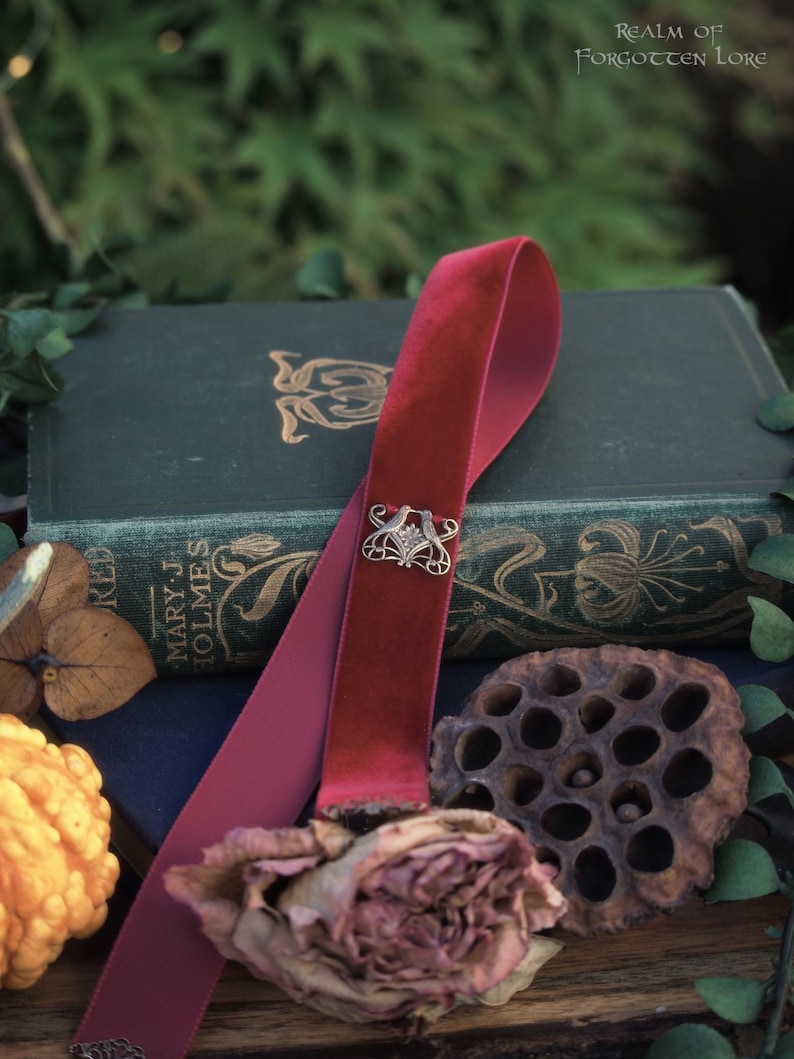 May include: A burgundy velvet bookmark with a silver filigree clasp rests on a vintage book. The bookmark is draped over the book, with a dried rose and other natural elements in the foreground. The text "Realm of Forgotten Lore" is visible.
