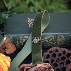 May include: A green velvet ribbon with an ornate silver clasp, draped over a stack of antique books. The ribbon is accented by dried flowers and seed pods, creating a vintage aesthetic. The books have gold lettering.