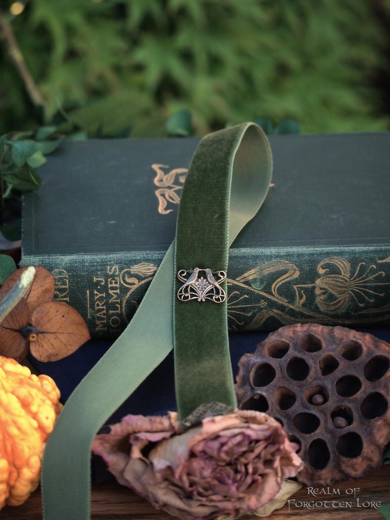 May include: A green velvet ribbon with an ornate silver clasp, draped over a stack of antique books. The ribbon is accented by dried flowers and seed pods, creating a vintage aesthetic. The books have gold lettering.