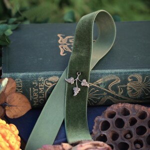 May include: A green velvet ribbon with a silver leaf charm rests on a dark green book. The book has gold lettering and floral designs. The text "Realm of Forgotten Lore" is visible. Dried flowers and a gourd are in the foreground.