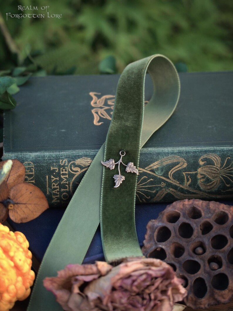 May include: A green velvet ribbon with a silver leaf charm rests on a dark green book. The book has gold lettering and floral designs. The text "Realm of Forgotten Lore" is visible. Dried flowers and a gourd are in the foreground.