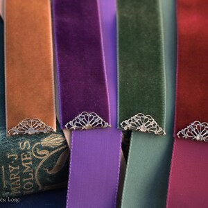 May include: Close-up of several velvet ribbon bookmarks in shades of orange, purple, green, and red. Each bookmark has a decorative silver-colored metal corner piece. The bookmarks are displayed on a vintage book.