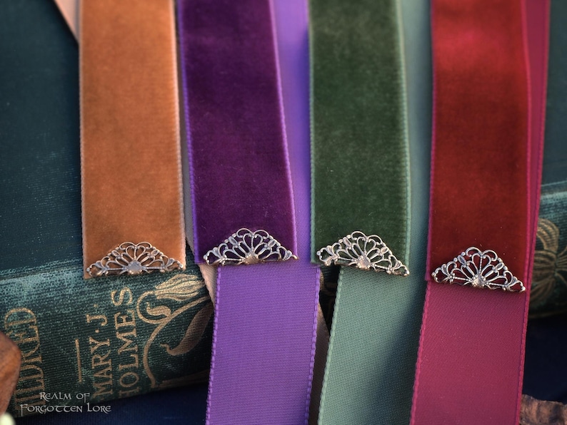 May include: Close-up of several velvet ribbon bookmarks in shades of orange, purple, green, and red. Each bookmark has a decorative silver-colored metal corner piece. The bookmarks are displayed on a vintage book.