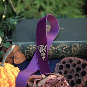 May include: A purple velvet ribbon bookmark with an ornate silver-colored metal accent. The bookmark is draped over a vintage book with gold lettering. A small gourd, dried rose, and seed pod are in the foreground.