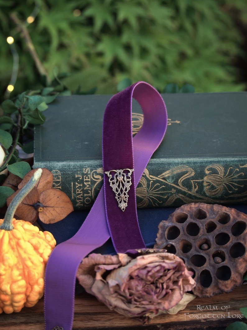 May include: A purple velvet ribbon bookmark with an ornate silver-colored metal accent. The bookmark is draped over a vintage book with gold lettering. A small gourd, dried rose, and seed pod are in the foreground.