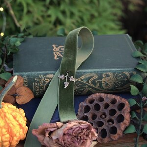 May include: A still life arrangement featuring a green velvet ribbon with a silver leaf charm, draped over a stack of antique books. A dried rose, a gourd, and a seed pod are also present. The text "Realm of Forgotten Lore" is visible.