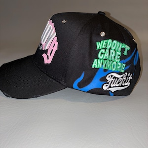 May include: A black baseball cap featuring a distressed brim and silver eyelets. The side displays blue flame graphics, the green embroidered text "WE DON'T CARE ANYMORE," and the white script text "Fuckit." The front features pink lettering.