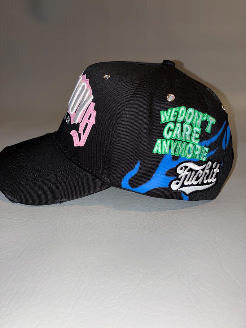 May include: A black baseball cap featuring a distressed brim and silver eyelets. The side displays blue flame graphics, the green embroidered text "WE DON'T CARE ANYMORE," and the white script text "Fuckit." The front features pink lettering.