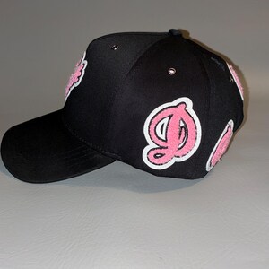 Puede incluir: Una gorra de b&eacute;isbol negra con parches de letras de felpilla rosa y blanca en los paneles laterales y frontales. La gorra incluye ojales met&aacute;licos en la corona y una visera curva, mostrando un dise&ntilde;o texturizado con letras estilizadas.