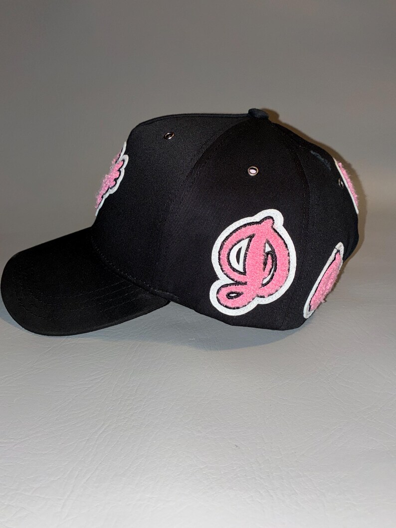 Puede incluir: Una gorra de b&eacute;isbol negra con parches de letras de felpilla rosa y blanca en los paneles laterales y frontales. La gorra incluye ojales met&aacute;licos en la corona y una visera curva, mostrando un dise&ntilde;o texturizado con letras estilizadas.