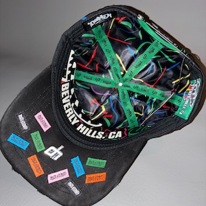 May include: A black baseball cap with a black and white chequered pattern on the inner brim and the text BEVERLY HILLS, CA. The interior lining is black with colourful abstract lines and green branded tape. The exterior features small colourful patches.