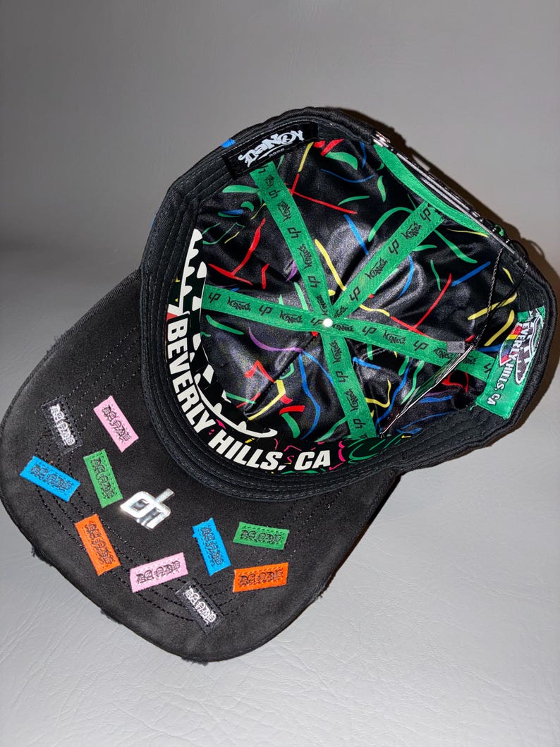 May include: A black baseball cap with a black and white chequered pattern on the inner brim and the text BEVERLY HILLS, CA. The interior lining is black with colourful abstract lines and green branded tape. The exterior features small colourful patches.