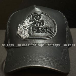 May include: Black trucker hat with a leather-like front panel and mesh back. The hat features an embroidered patch with a fish skeleton and the text "YO NO PESCO" in white and black.