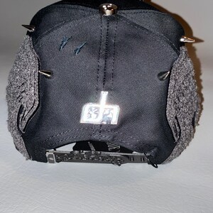 May include: A black baseball cap featuring silver metal spikes on the crown, grey textured side panels, and a metallic silver logo emblem on the back. The cap includes an adjustable black strap with a detailed buckle and a small black tag.