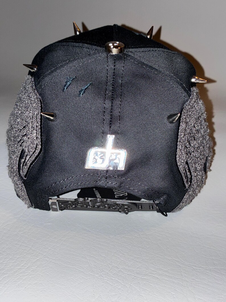 May include: A black baseball cap featuring silver metal spikes on the crown, grey textured side panels, and a metallic silver logo emblem on the back. The cap includes an adjustable black strap with a detailed buckle and a small black tag.