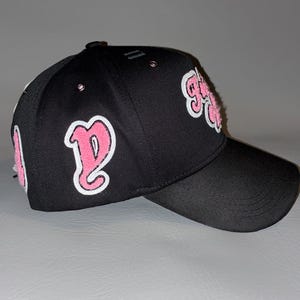 Puede incluir: Una gorra de b&eacute;isbol negra con parches bordados en rosa y blanco en los paneles laterales y frontales. La gorra tiene una corona estructurada, una visera curva y peque&ntilde;os ojales met&aacute;licos en los paneles superiores. Los parches muestran letras estilizadas.
