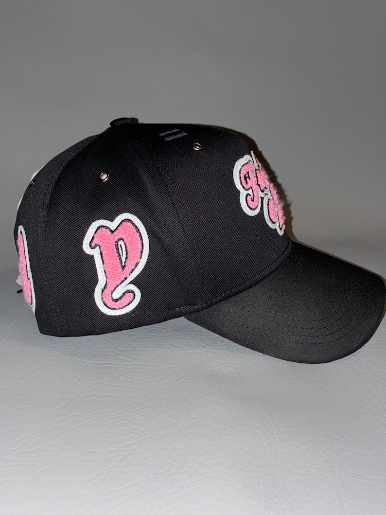 Puede incluir: Una gorra de b&eacute;isbol negra con parches bordados en rosa y blanco en los paneles laterales y frontales. La gorra tiene una corona estructurada, una visera curva y peque&ntilde;os ojales met&aacute;licos en los paneles superiores. Los parches muestran letras estilizadas.