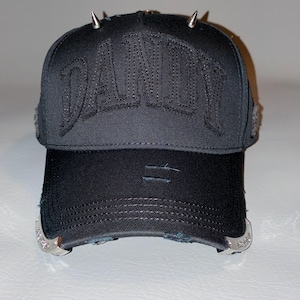 May include: A black baseball cap featuring the word DANDY embroidered in black on the front panel. The cap includes silver-toned metal spikes on the top, distressed detailing on the brim, and decorative silver-toned metal accents on the sides.