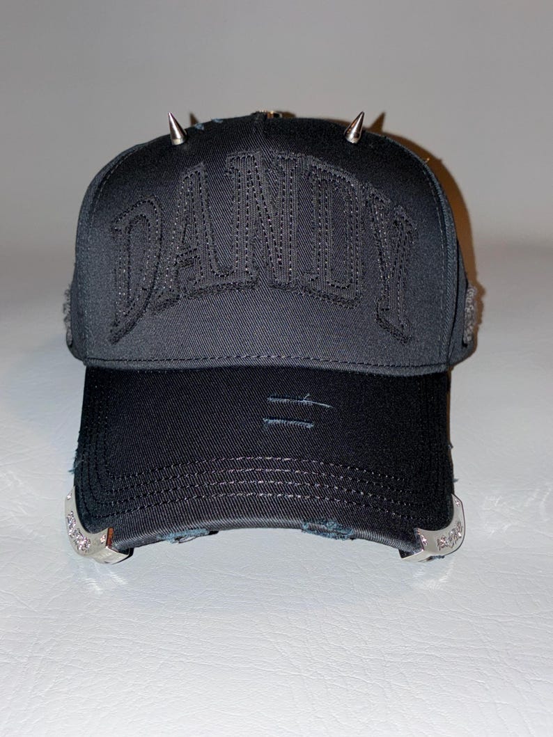 May include: A black baseball cap featuring the word DANDY embroidered in black on the front panel. The cap includes silver-toned metal spikes on the top, distressed detailing on the brim, and decorative silver-toned metal accents on the sides.