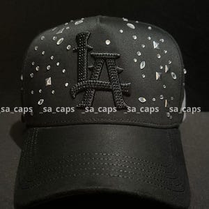 May include: Black baseball cap with the letters "LA" in a raised, textured design. The cap is embellished with scattered, sparkling rhinestones. The brim is curved, and the cap has a structured fit.