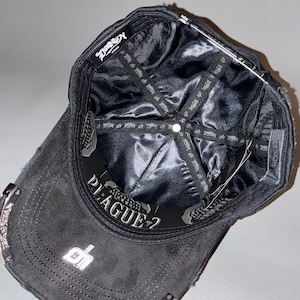 May include: A black baseball cap shown from an interior perspective, featuring a shiny black satin lining with repeating text. The inner band displays the word PLAGUE-? in white lettering, and a silver metallic logo is visible on the brim.