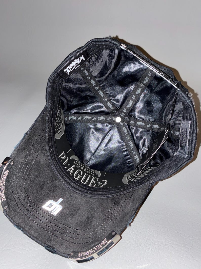 May include: A black baseball cap shown from an interior perspective, featuring a shiny black satin lining with repeating text. The inner band displays the word PLAGUE-? in white lettering, and a silver metallic logo is visible on the brim.