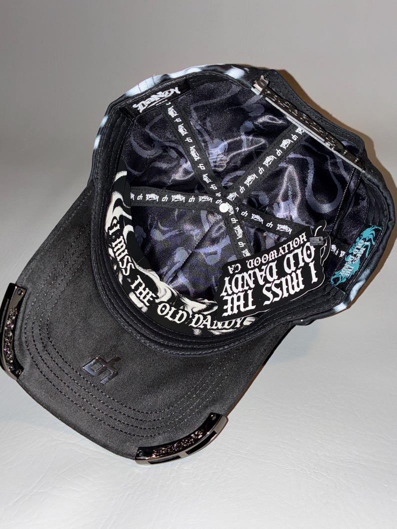 Puede incluir: Una gorra de b&eacute;isbol negra vista desde el interior, con un forro de sat&eacute;n negro con un patr&oacute;n de logotipo blanco repetitivo. La banda interior para el sudor muestra el texto I MISS THE OLD DANDY HOLLYWOOD, CA en letras g&oacute;ticas blancas.