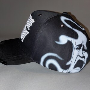 Puede incluir: Una gorra de b&eacute;isbol negra con texto bordado en blanco en el lateral y un gr&aacute;fico de aer&oacute;grafo blanco de una cara llorando en el panel posterior. La gorra incluye ojales met&aacute;licos en la corona y una peque&ntilde;a placa met&aacute;lica en la visera curva.