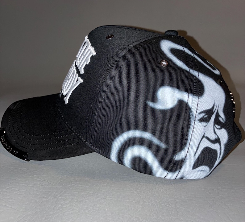 Puede incluir: Una gorra de b&eacute;isbol negra con texto bordado en blanco en el lateral y un gr&aacute;fico de aer&oacute;grafo blanco de una cara llorando en el panel posterior. La gorra incluye ojales met&aacute;licos en la corona y una peque&ntilde;a placa met&aacute;lica en la visera curva.