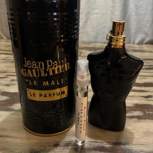 Puede incluir: Una botella de perfume negra con forma de torso, un recipiente cilíndrico y un vial pequeño. El recipiente cilíndrico y la botella con forma de torso tienen detalles dorados. El texto "Jean Paul Gaultier Le Male Le Parfum" es visible.