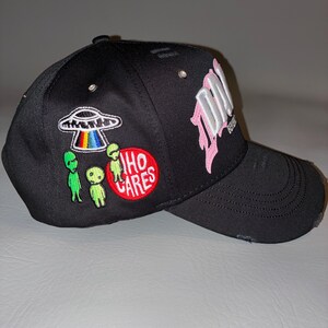 May include: A black baseball cap featuring distressed detailing on the brim and crown. The side displays embroidered patches of a UFO with a rainbow beam, three green alien figures, and a red circular patch with the white text WHO CARES.